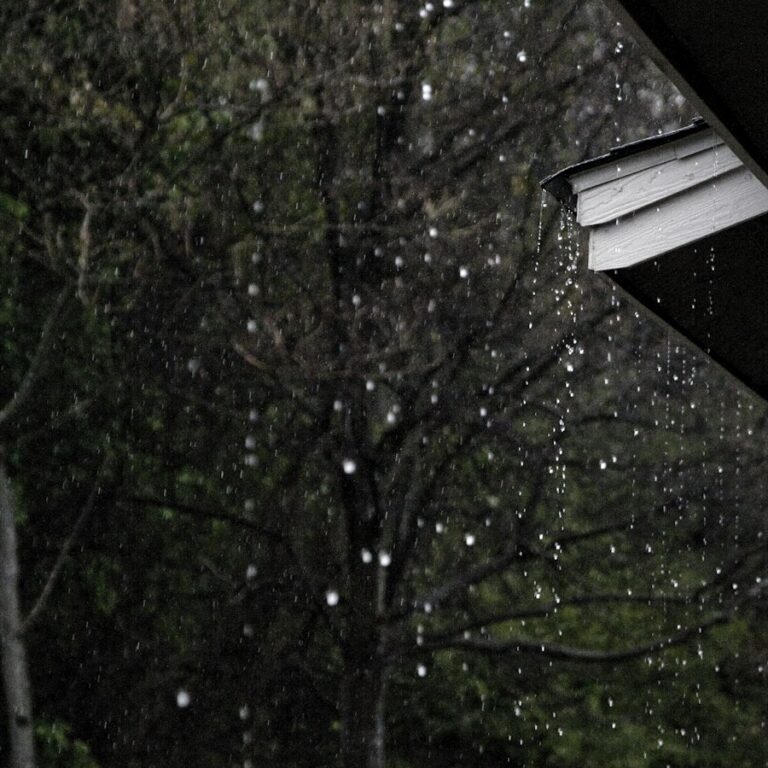 Regen tropft vom Dachvorsprung eines Hauses vor grünen Bäumen
